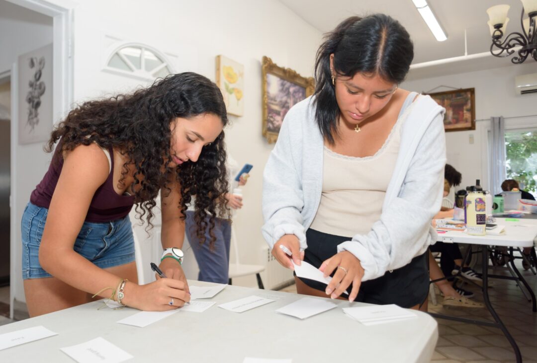 Two girls write on note cards, leaning over a white table with other cards to be filled out. There's another table in the background with more participants and materials.
