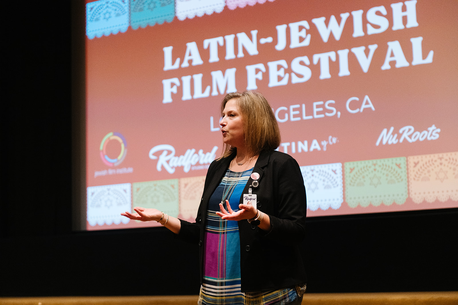 A woman presenting in front of a large projected screen which reads, "Latin Jewish Film Festival" with various participating organizations' logos along the bottom.